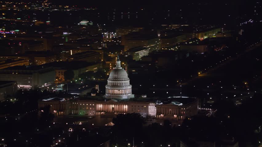 Spectacular aerial drone shot of the illuminated United States Capitol building at night. The high-angle perspective looks straight down the National Mall, capturing the glowing Washington Monument in the distance, surrounded by Washington D.C. city lights and street traffic.