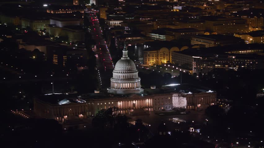 Stunning nighttime aerial drone shot of the brightly illuminated United States Capitol Building and surrounding city streets in Washington D.C., showcasing the iconic dome and classic architecture from above.