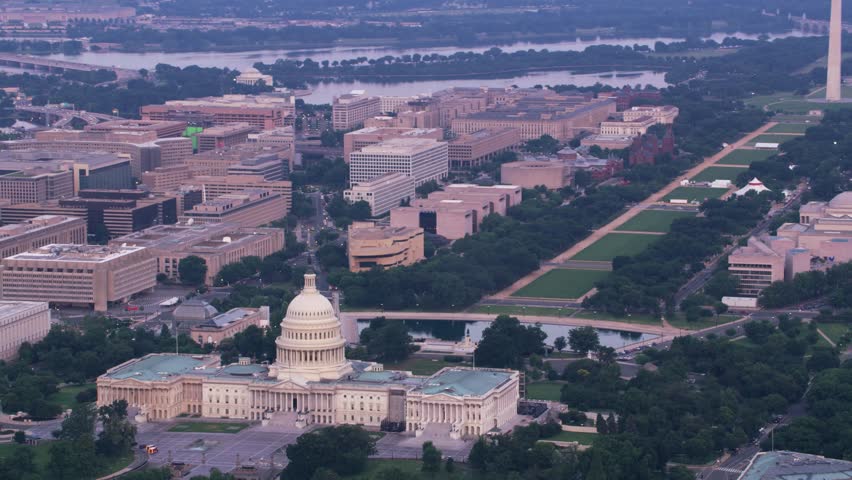 Drone footage of Washington, D.C. showcasing the United States Capitol building with its iconic dome and neoclassical architecture. The aerial view extends across the National Mall, highlighting landmarks such as the Washington Monument, government buildings, and museums arranged in a grid-like pattern. The Potomac River is visible in the background, adding geographic context to this historic and political center of the United States.