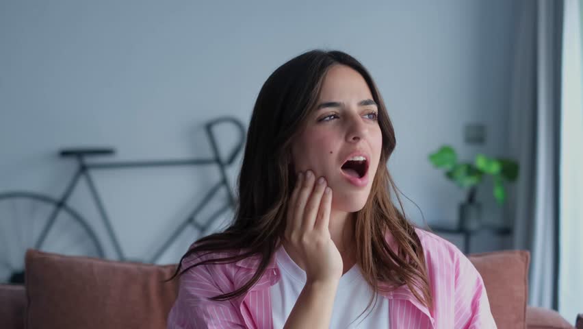 A young woman is visibly suffering from severe tooth pain and jaw discomfort while sitting on a couch in her modern home. She touches her cheek, seeking relief from the agonizing sensation.