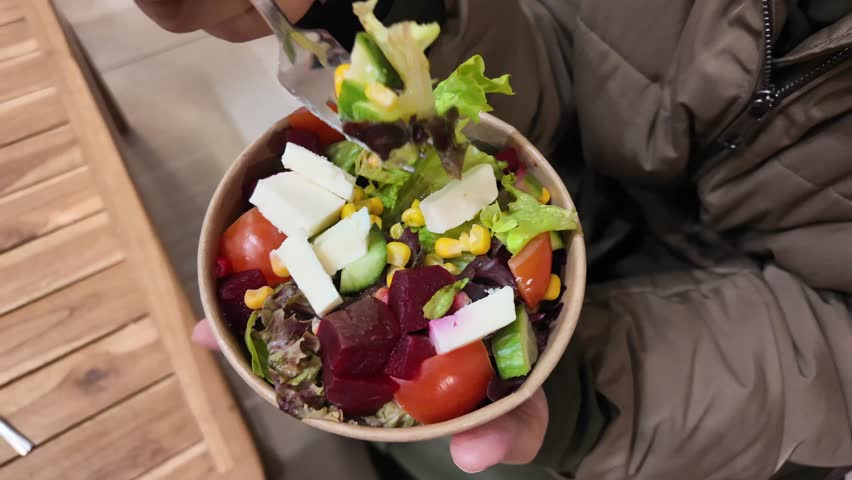 A person is mixing a colorful healthy salad with a fork in a paper bowl while sitting at a wooden table, capturing the dynamic motion of food preparation and healthy eating