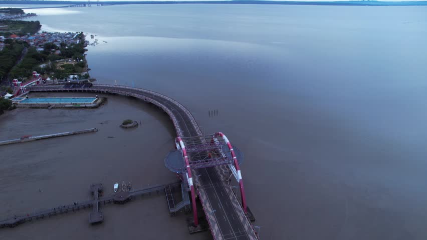 Aerial View of Suroboyo Bridge at Dawn: A high-angle drone shot capturing the iconic red-arched Suroboyo Bridge stretching across the coastal area of Kenjeran, Surabaya, Indonesia, during a calm, overcast morning
