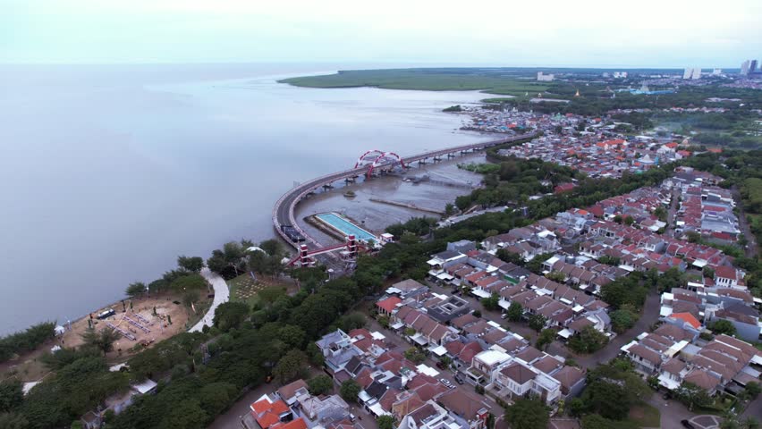 Aerial view of the iconic Suroboyo Bridge in Surabaya, Indonesia, showing its unique curved design and red structural arches spanning the coastal area of Kenjeran