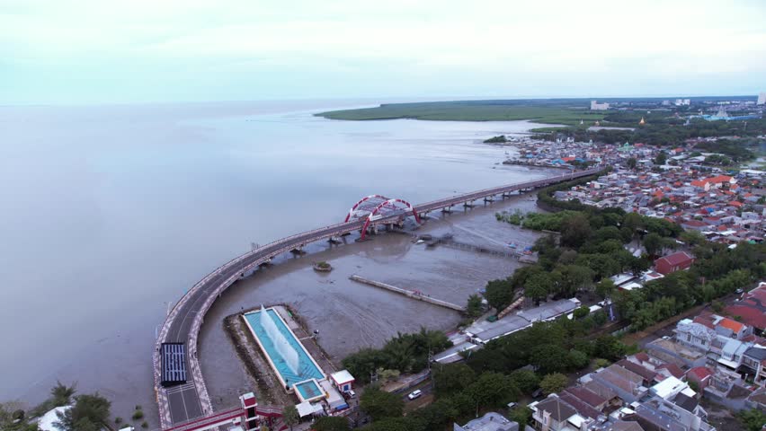 The modern Suroboyo Bridge connecting the coastal tourism areas of Kenjeran, Surabaya, captured from an aerial perspective under a soft, overcast morning sky