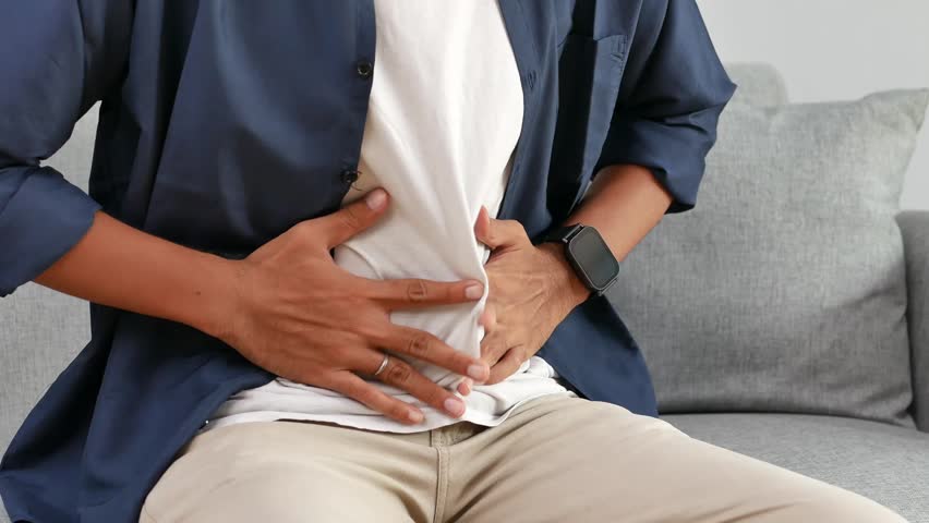 A man in a casual clutches his abdomen in discomfort, experiencing stomach pain or indigestion, captured in a close-up sitting on sofa at home