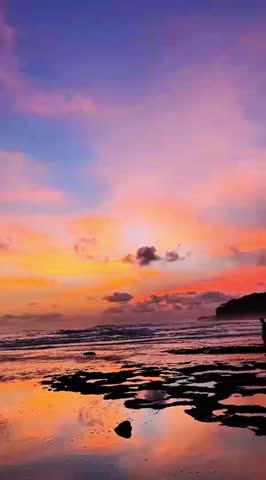 A stunning purple sunset bathes a rocky beach, casting vibrant reflections in the water. Dramatic, dramatic clouds paint the sky above the crashing waves and dark cliffs."