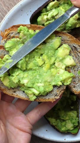 Close up view of a hand holding a slice of bread and spreading fresh, mashed avocado on it with a knife. Healthy eating concept for a nutritious breakfast or a wholesome vegan snack