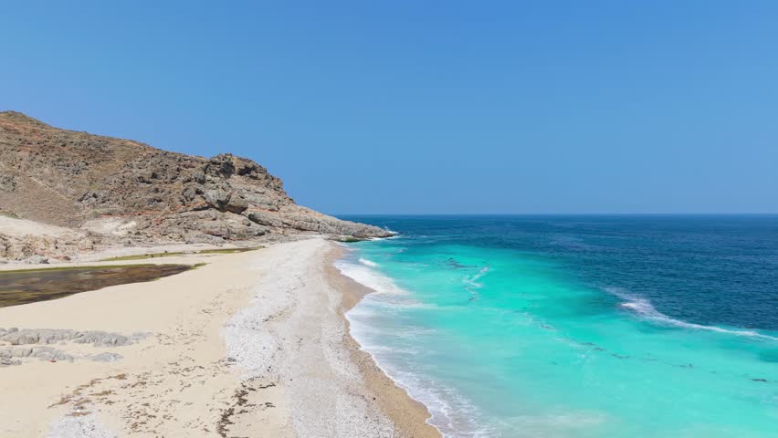 Aerial view of turquoise sea water meeting a white sand beach and rugged mountain cliffs along the coastline of Saudi Arabia.
