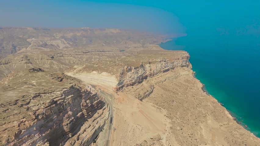 Aerial view of steep limestone cliffs and a winding asphalt road along the turquoise coastline of the Arabian Sea in Dhofar, Oman.