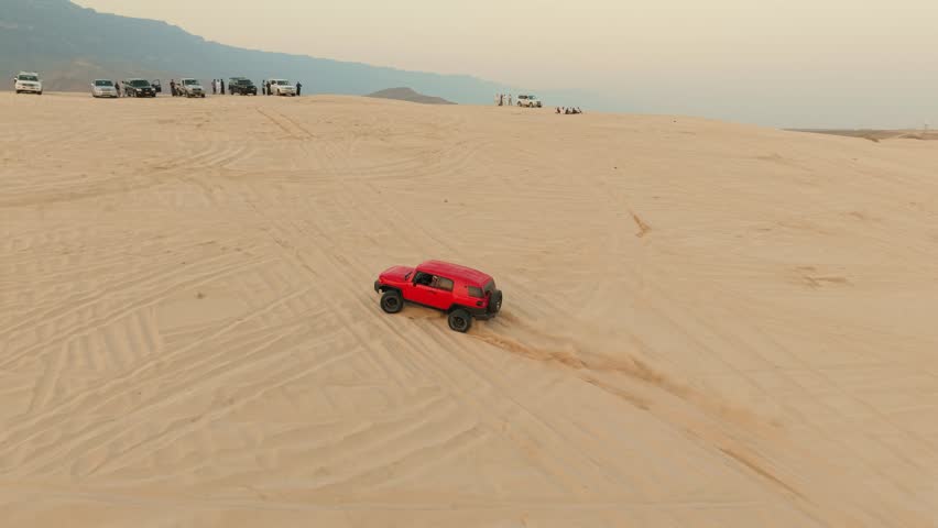 Aerial view of a red SUV driving through sand dunes with men and parked vehicles nearby in the desert of Salalah, Oman.