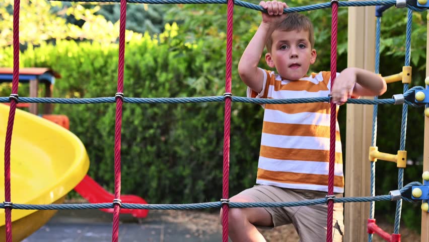 A 5-6-year-old boy climbs a rope ladder at a park playground in the summer. Harmonious physical development of a child