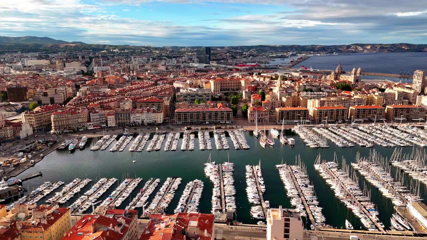 Panoramic drone view of Marseille’s Vieux-Port marina in France, with rows of yachts, historic waterfront buildings, ferry terminals and the city skyline stretching toward the hills.