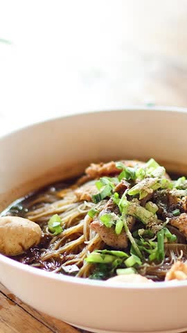 A close-up vertical shot of a hand sprinkling spicy chili flakes onto a noodle dish.
