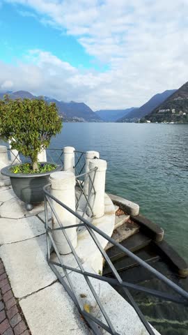 Beautiful view of Como lake, Italy. Promenade with green trees in Como town. Clouds over the Alps mountains. Beautiful landscape at sunny day. Vertical video
