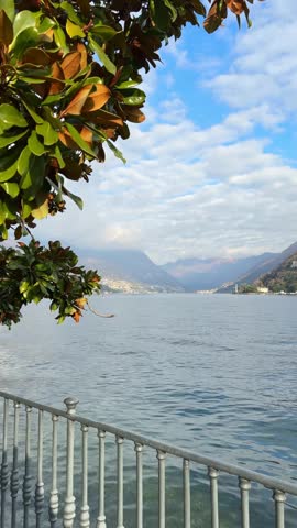 Beautiful view of Como lake, Italy. Promenade with green trees in Como town. Clouds over the Alps mountains. Beautiful landscape at sunny day. Vertical video

