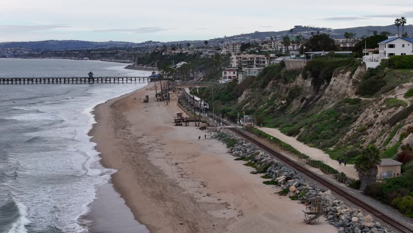 Aerial view of an Amtrak train traveling beside a sandy beach and coastal cliffs near a pier, with ocean waves, hillside homes, and shoreline scenery in Southern California.