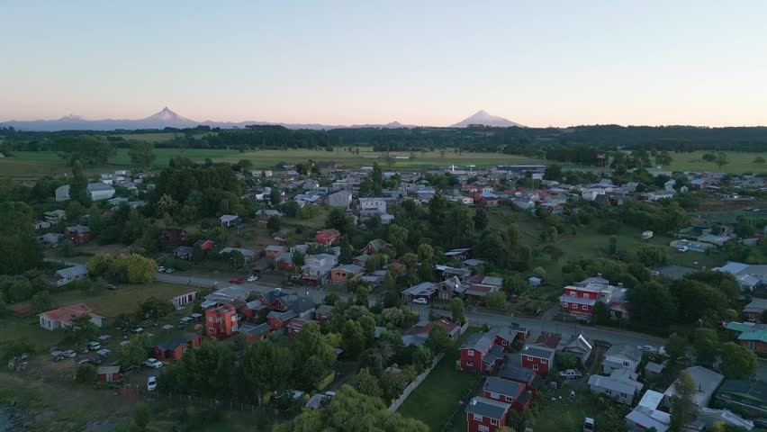 Aerial view of small southern Chilean town surrounded by farmland and forest with snow-capped Andean volcanoes on the horizon at sunset. Entre Lagos, Chile