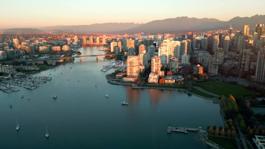 Aerial drone view of downtown Vancouver over False Creek at golden hour, featuring waterfront high rises, marina, bridge, mountains, and the city skyline in British Columbia, Canada.