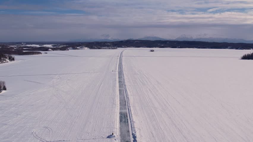 Aerial 4K 60fps drone footage over frozen Big Lake, Alaska. Carved ice road leads to distant snow-capped mountains through clouds and haze across vast snow-covered lake.