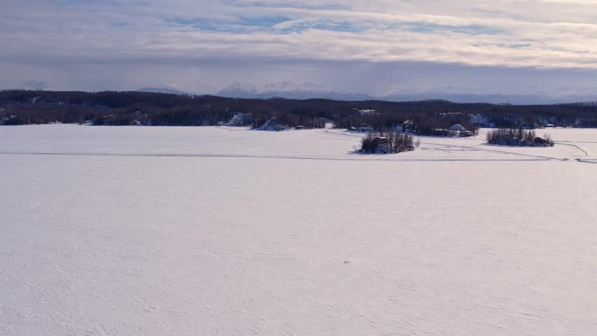 Aerial 4K 60fps drone footage over frozen Big Lake, Alaska. Ice road crosses frame horizontally, island right of center, lakeside community and snow-capped mountains line distant shore.