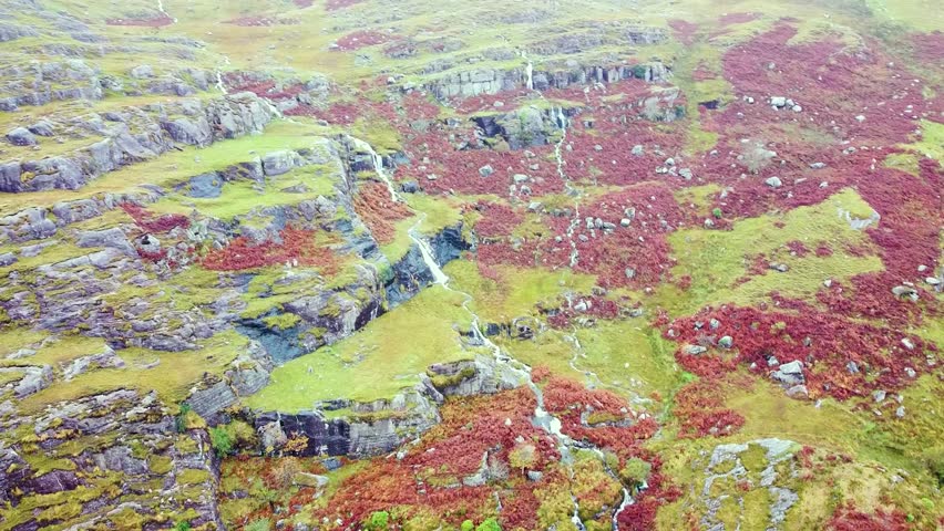 Drone shot capturing a rugged rocky outcrop and a small cascading waterfall surrounded by lush green grass and vibrant purple autumn heather of the Western Irish mountains on a foggy day