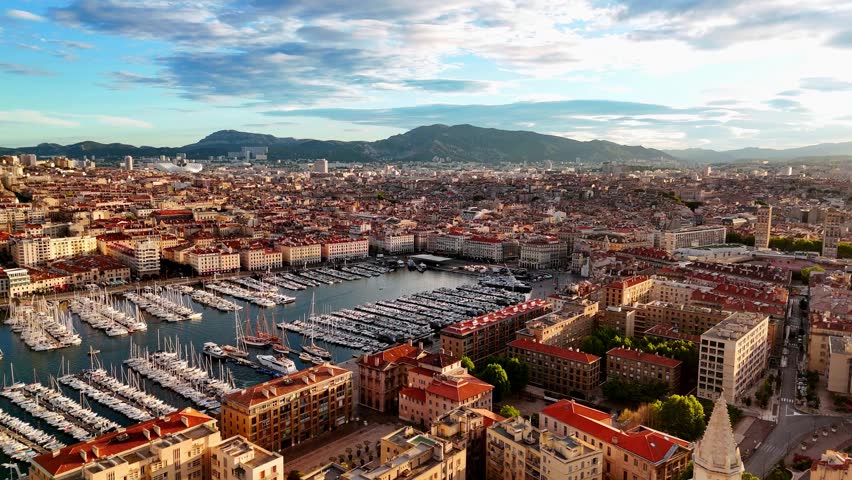Aerial panorama of Marseille, France, overlooking the Vieux-Port marina filled with yachts, historic rooftops, and distant hills in warm evening light beneath a dramatic sky.