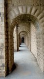 Perspective view of a long, vaulted stone corridor featuring a series of repetitive, hand-carved arches. The video showcases the intricate masonry and weathered texture of the sandstone or limestone blocks, creating a rhythmic architectural pattern. Soft daylight filters into the passage, highlighting the depth and symmetry of the historic structure. This atmospheric footage captures the essence of ancient Mediterranean or Middle Eastern architecture, ideal for projects focused on heritage, travel, historical storytelling, or architectural design. - Powered by Shutterstock - Get 15% off with code: PIKWIZARD15