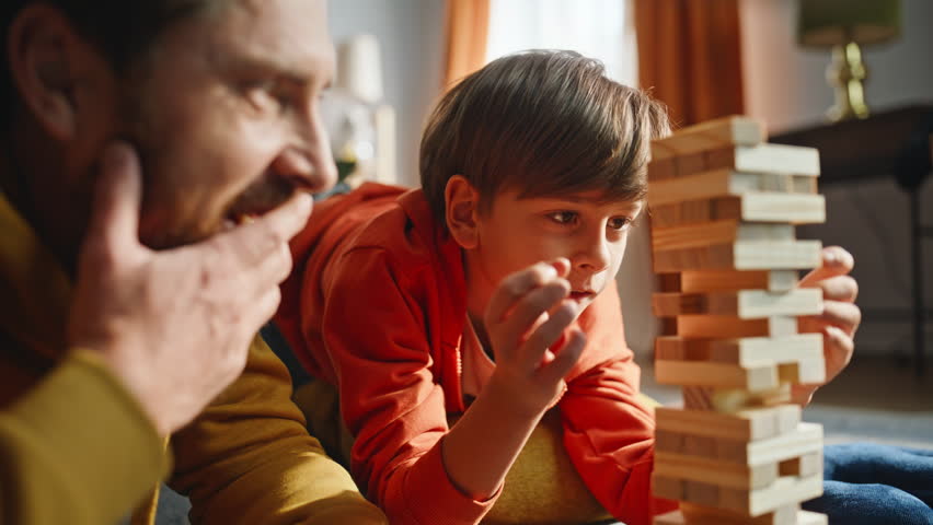Man son playing board game laughing spending family leisure lying floor closeup. Focused boy taking wooden block from towel holding balance with concentration. Smiling father child involved in game.