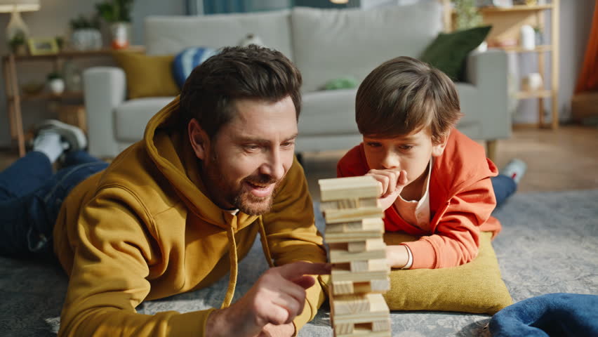Father son playing game lying apartment floor closeup. Smiling bearded man playful preteen boy building tower wooden blocks enjoying family board game together. Happy parent child togetherness home