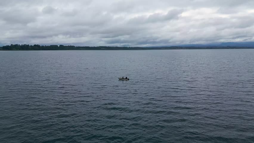 Aerial tracking shot following two people on a small boat crossing a wide gray lake with forested shoreline under dramatic overcast sky