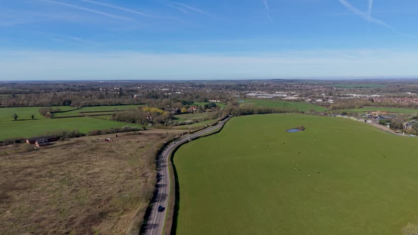 London Road near Hertford Heath, Hertfordshire, England. Left pan across rural fields and hedgerows under clear blue spring sky with light traffic on a quiet country lane.
