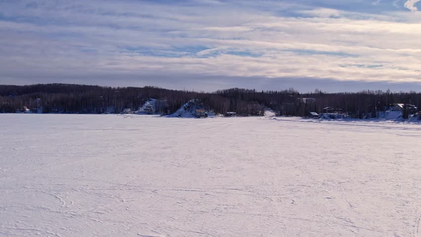 Aerial 4K 60fps drone footage flying toward shore over frozen Big Lake, Alaska. Vast snow-covered ice leads to lakeside homes and dense treeline, sun breaking through layered clouds.