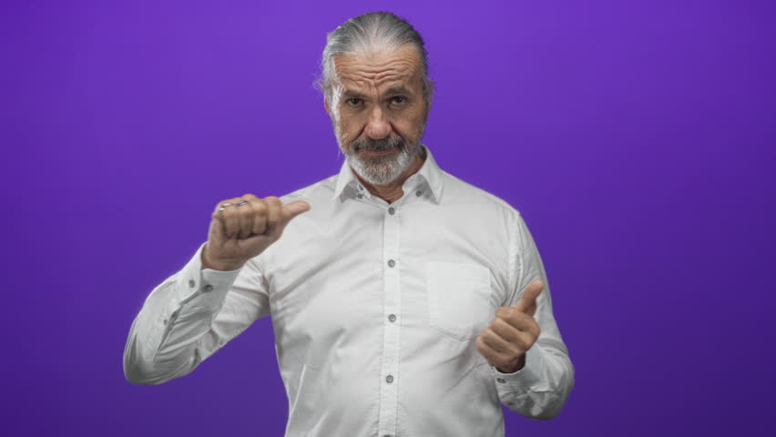 Man points thumbs to chest in purple studio set, hoary middle age with long gray hair and beard, wearing white shirt and rings on fingers; confidence.