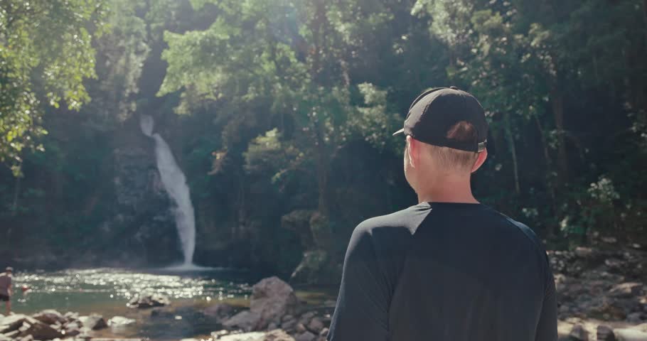 Man standing and looking at tropical waterfall in lush green jungle forest. Back view of traveler exploring nature during sunny day. Adventure and outdoor lifestyle.