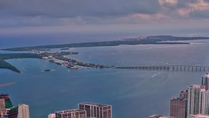 The shoreline of Miami is visible with buildings and boats along the water. Islands dot the sea in the distance under a cloudy sky. The view shows the diverse layout of the area.