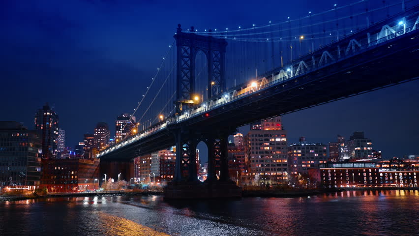 Manhattan Bridge structure over East River at night in NYC. Wide angle night shot of the Manhattan Bridge with illuminated buildings and water reflections.