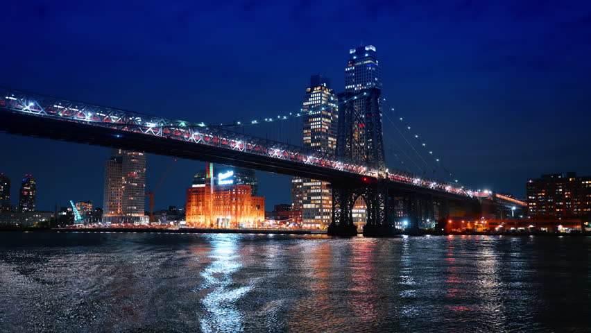 Brooklyn Williamsburg waterfront and bridge at night with lights. View of the Domino Sugar Refinery area and Williamsburg Bridge illuminated at night on East River.