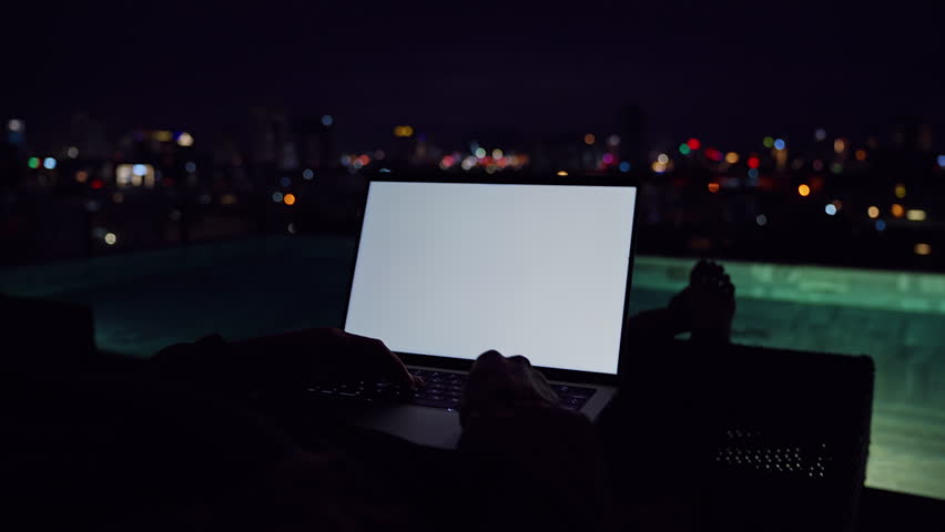 Woman Using Laptop with Blank Screen Mockup on Rooftop Near Pool Overlooking Night City Lights in a Low Angle Dark View.