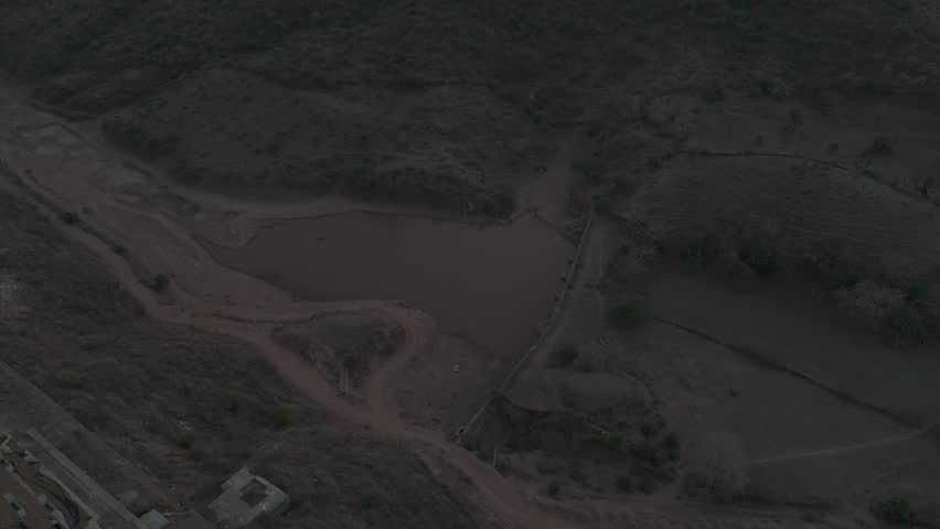 Top-down aerial view of a pond near the Aravalli hills around Udaipur, Rajasthan, showing calm water surrounded by rugged terrain, native trees, and rural landscape.