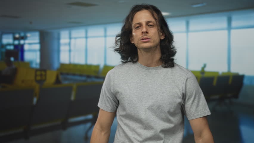 Man with long hair wearing gray t shirt, arms outstretched and looking sideways in airport terminal near yellow seats; travel delay anticipation.