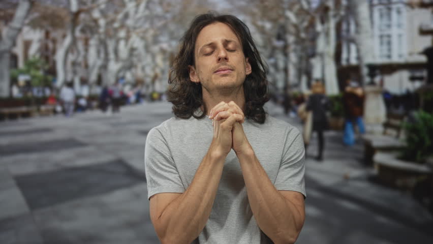 Man with clasped hands and pleading expression on a crowded street with benches and trees, long hair and bare forearms visible; pleading hope.