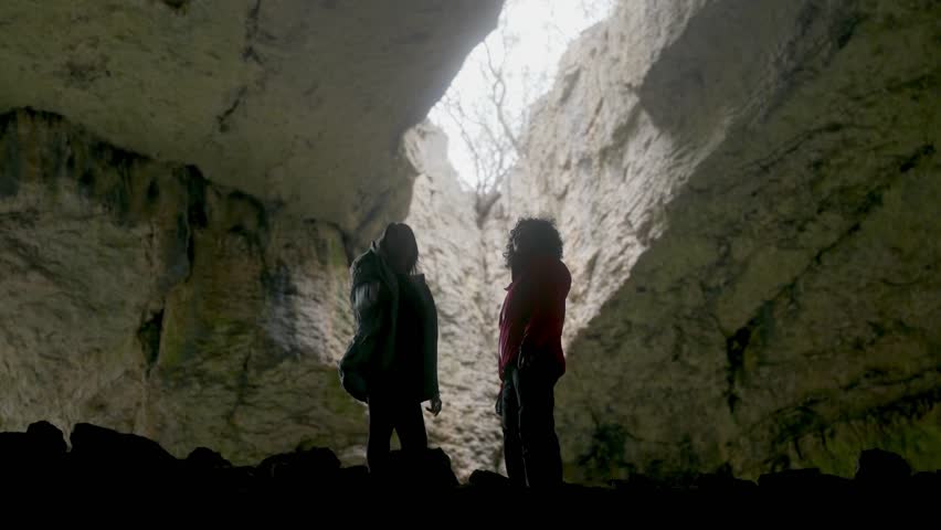 Silhouetted figures of two women exploring the vast, natural beauty of a majestic cave. One points towards the inspiring light, sharing a moment of connection and discovery in this ancient, awe-inspiring natural wonder.-