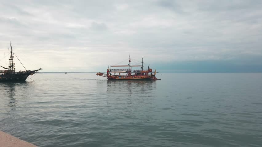 A picturesque vintage wooden tourist boat, resembling a pirate ship, cruises gently across the serene waters of Thessaloniki Bay, Greece, under a soft, cloudy sky.-