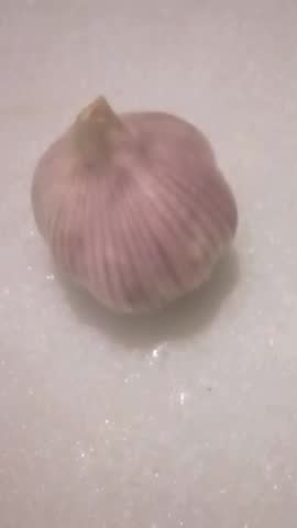 A high-angle, close-up shot of a fresh, whole garlic bulb with distinctive purple stripes on its skin. The garlic is placed on a white, slightly reflective surface, making it ideal for culinary, health, and food-related content.