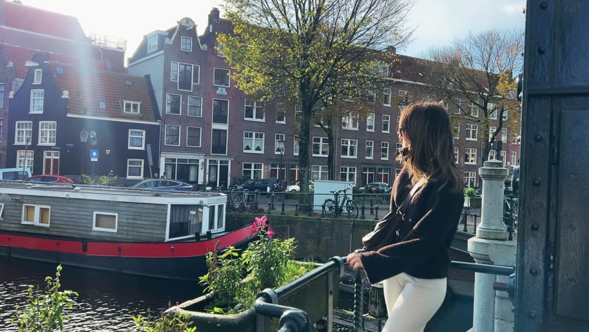 A young woman stands on a bridge, looking at traditional canal houses, a houseboat, and a canal in Amsterdam on a sunny day. This authentic moment captures urban exploration and travel.