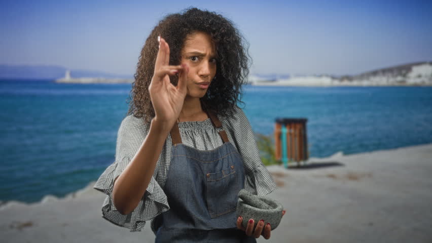 Woman holding mortar and pointing finger near coastal building while wearing apron and denim; curiosity teaching skill.