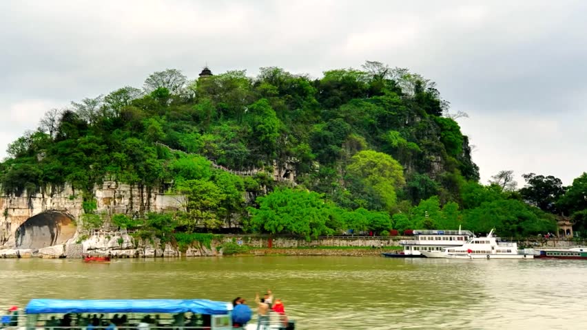 A vibrant, green hill rises dramatically from the Li River, adorned with trees and featuring visible cave entrances. Structures and stairs are carved into the hillside, with boats visible on the water. high-quality professional detailed modern elegant stylish clean crisp