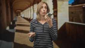 Woman speaking into smartphone with open palm questioning gesture in sunlit colonnade building; concern communication. - Powered by Shutterstock - Get 15% off with code: PIKWIZARD15
