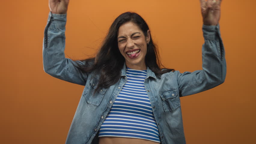 Woman wearing denim shirt and striped crop top showing rock horns with both hands and tongue out in orange studio; playful rebellion.