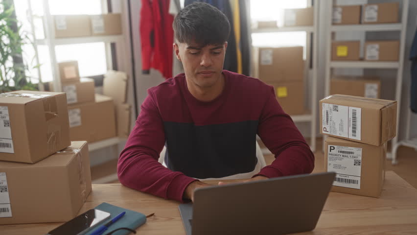 Man smiling at laptop with stacked cardboard boxes on desk in a building shipping room; entrepreneur pride.